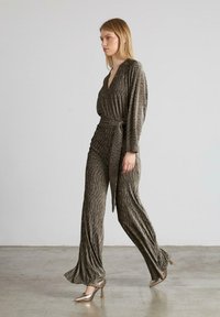Mono verde olivo con escote en V profundo, mangas anchas, tela texturizada y cintura con cinturón; combinado con tacones metálicos de punta.