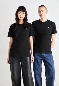 Camisetas de algodón negras de manga corta, con un pequeño logo bordado en el lado izquierdo del pecho, combinadas con pantalones de cuero negros y jeans azules.