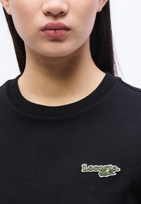 Κοντινό πλάνο ενός ατόμου που φοράει μαύρη μπλούζα με ένα μικρό πράσινο λογότυπο Lacoste στο στήθος.