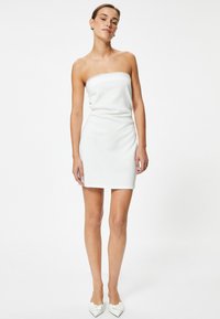 Vestido blanco sin mangas con una silueta ajustada, textura suave y detalles de encaje en el escote; combinado con zapatos blancos de punta fina.