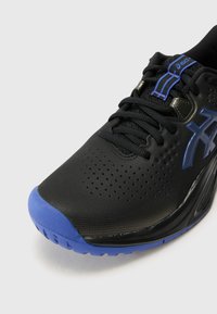 Chaussure de sport noire avec une tige texturée, des accents bleus sur le bout et le logo, et un système de fermeture à lacets. Elle comprend des perforations pour la respirabilité.