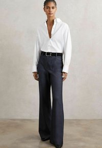 Cămașă albă cu guler în V adânc și mâneci lungi, asortată cu pantaloni negri, largi și un curea neagră cu o cataramă argintie.