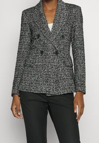 Dubbelgeknoopte blazer van een gestructureerde zwart-witte stof, voorzien van grote zwarte knopen en twee voorzakken. Gecombineerd met een zwarte broek.