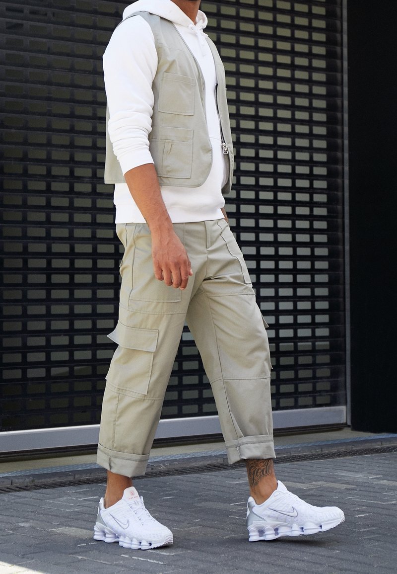 Homme marchant en extérieur portant un sweat à capuche blanc, un gilet utilitaire beige et un pantalon cargo, des baskets blanches Nike, avec un tatouage à la cheville visible sur la jambe droite.