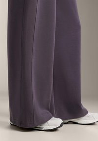 Široké tmavě fialové kalhoty vyrobené z měkké tkaniny s proplétanými bočními panely, doplněné o bílé sportovní boty s černými podrážkami.