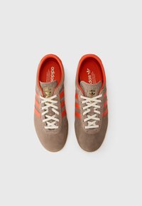Hnědé semišové tenisky se třemi oranžovými pruhy, bílými tkaničkami a béžovou gumovou podrážkou. Červený vnitřek s zlatým logem adidas na jazyku.
