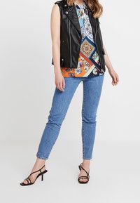 Gilet senza maniche in pelle nera sopra una blusa a fantasia colorata, abbinato a jeans skinny blu e tacchi neri a strappo.