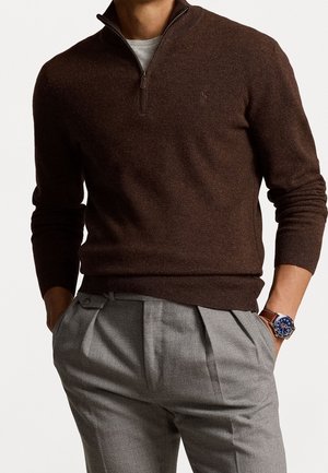 Homme portant un pull marron à demi-fermeture éclair par-dessus une chemise grise, un pantalon gris plissé et une montre à bracelet en cuir marron avec un cadran bleu.