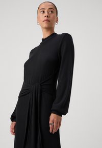 Črna rebrasta obleka z dolgimi rokavi, visokim ovratnikom in zavezanjem v pasu. Gladka tekstura in ohlapen silhuet. Srebrni prstan na roki.
