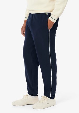 Pantalones de chándal azul marino en tejido suave, con bolsillos laterales y una franja blanca con la marca que recorre la pierna. Con un logotipo como acento.