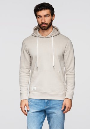 Mężczyzna z brodą, ubrany w jasnobeżową bluzę z kapturem, z kieszenią z przodu i sznurkami, w połączeniu z niebieskimi dżinsami, stoi na jednolitym tle.