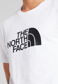 Valge puuvillane T-särk, mille esiküljel on suur must graafiklogo "THE NORTH FACE", kaarjas disain ja julge kiri.