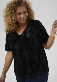 Donna sorridente con capelli biondi ricci che indossa un top nero di velluto a V, jeans blu e un bracciale di perle sul braccio destro sollevato.