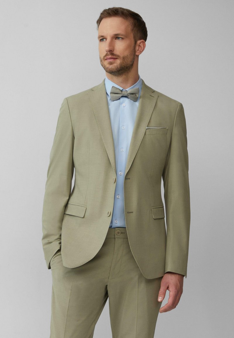 Costume vert clair avec revers crantés, fermeture à un bouton, et pantalon assorti. Associé à une chemise bleu pâle à boutons et un nœud papillon.