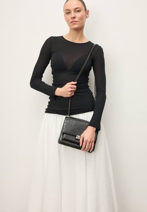 Schwarzes Langarm-Oberteil mit transparentem Ausschnitt, kombiniert mit einem weißen, fließenden Rock. Das Model trägt eine kleine schwarze Tasche mit Krokodilstruktur.