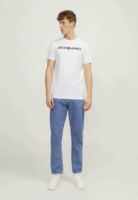 White cotton t-shirt with black and red "JACK & JONES" logo, paired with light blue denim jeans and white sneakers. Fit is casual.