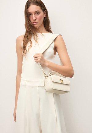 Naine kannab valget varrukateta toppi ja laiade jalgadega valgeid pükse, hoides käes kreemjat ristikandmist kulla klambri jaotusega.