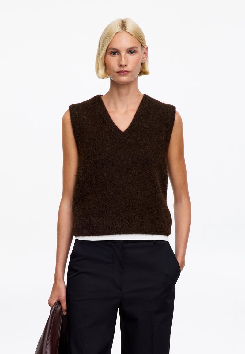 Femme blonde portant un gilet sans manches en fourrure marron foncé sur une chemise blanche et un pantalon noir, tenant un sac en cuir marron dans sa main gauche.