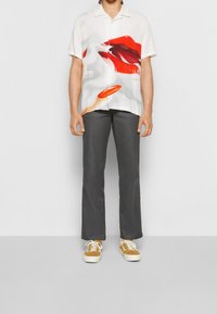 Valkoinen lyhythihainen paita, jossa on rohkea punainen huuligrafiikka, yhdistettynä harmaisiin suoriin housuihin ja keltaisiin tennareihin, joissa on valkoiset pohjat.