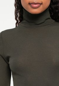 Kobieta z kręconymi włosami w oliwkowym swetrze z prążkowanym kołnierzem, widoczna od ramion do warg na białym tle.