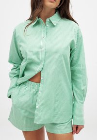 Zaļi un balti svītrota pogājamā krekla ar brīvu piegriezumu komplekts ar atbilstošām šortiem. Gluda auduma un garas piedurknes ar pogu aprocēm.