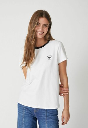Valge puuvillane T-särk, millel on must krae, lühikesed varrukad ja väike must logo rinnal, koos siniste denim-pükstega.