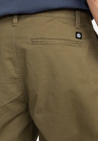Olive green cotton pants with a back pocket featuring a black logo patch. The fabric has a smooth texture and a classic design.