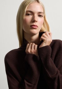 Pull en maille côtelée brun foncé avec un col montant, présentant des manches longues et une finition texturée. Gros plan de mains tenant le col.