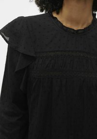 Blusa nera con colletto a volant, texture a pois, dettagli in pizzo e tessuto arricciato. Il materiale è leggero e trasparente.