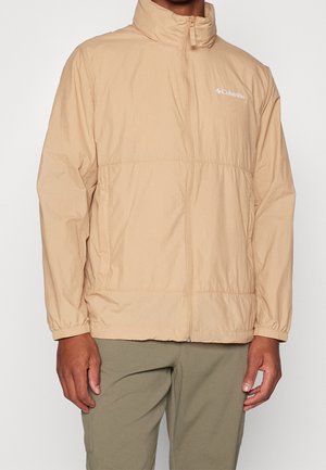 Veste beige Columbia à fermeture éclair avec poignets élastiques portée par une personne debout, associée à un pantalon vert olive, sur un fond uni.