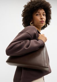 Bolsa de mão castanho em couro com forma triangular, textura suave e branding mínimo. A portadora usa um sweatshirt castanho escuro.