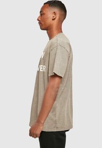 Lys brun, oversized T-shirt lavet af blødt, tekstureret stof med korte ærmer og rund halsudskæring. Stor hvid tekst på forsiden.