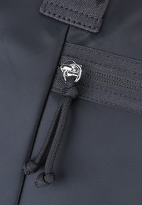 Podrobnost črne tkanine torbice, ki prikazuje zadrgo s srebrnim kovinskim obročkom in črnimi vrvicami. Material ima teksturiran in trpežen videz.