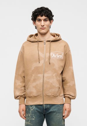 Mladý muž nosí béžovú mikinu na zips s logom "Aries" na hrudi a modré rifle s grafickými vzormi, stojí pred jednoduchým pozadím.