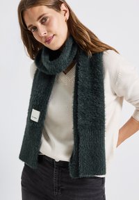 Mørkegrøn, fuzzy strikket tørklæde med rektangulær form og et mærke. Båret over en lys beige sweater, hvilket skaber et hyggeligt lag-på-lag look.