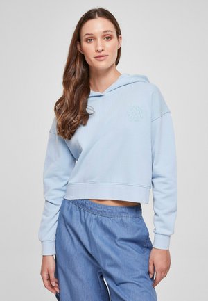 Lyseblå cropped hettegenser laget av mykt stoff, med en frontlomme og brodert blomstdetalj. Kombinert med avslappede blå bukser.