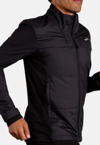 Giacca da corsa nera con zip frontale, colletto alto e pannelli opachi a contrasto; materiale leggero con una texture liscia e senza motivi.