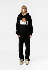 Sudadera negra con estampado gráfico y diseño oversize, combinada con pantalones negros y zapatillas multicolores. Elementos destacados: textura y detalle.