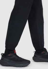 Personne portant des baskets de sport noires avec des détails rouges et un pantalon noir avec des poignets élastiques sur un fond clair uni.
