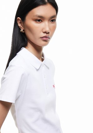 Nuori nainen, jolla on pitkät mustat hiukset ja valkoinen kauluksellinen poolopaita, jossa pieni punainen kirjottu logo, katsoo vakavana sivulle.