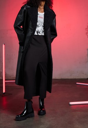 Manteau noir en similicuir avec un col en shearling noir texturé, associé à un t-shirt blanc à motif graphique et un pantalon large noir. Bottes noires épaisses.