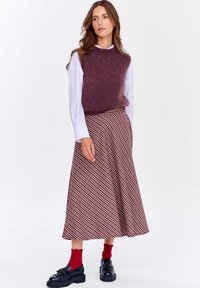 Žena v purpurových bezrukávových svetrích cez bielu košeľu, vzorovaných midi sukni, červených ponožkách a čiernych mokasínach na bielom pozadí.