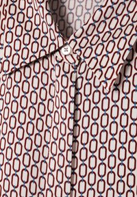 Rotes und blaues geometrisches Kettenmuster auf einem cremefarbenen Stoffhemd. Verfügt über einen spitzen Kragen und einen einzelnen Knopf am Ausschnitt.