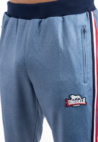 Lonsdale London bikses gaiši ziliem audumiem ar teksturētu apdari. Iezīmējas ar sarkaniem un baltiem sānu svītrām un logotipa uzšuvumu uz augšstilba.