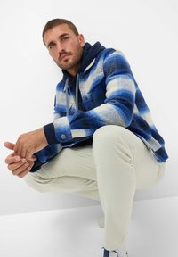 Giacca in pile a scacchi blu e bianchi con cappuccio, abbinata a pantaloni in corduroy color crema. La giacca presenta bottoni a pressione e un design testurizzato.