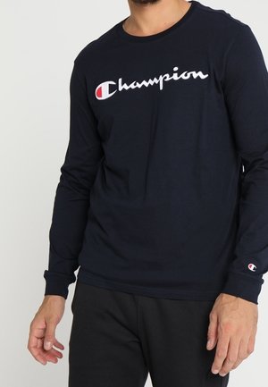 Mann trägt ein marineblaues Langarmshirt von Champion mit Logo über der Brust und kleinem Logo am linken Ärmelsaum, kombiniert mit schwarzen Hosen.