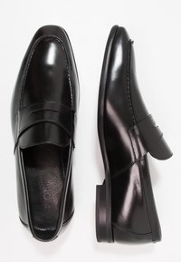 Paire de mocassins habillés en cuir noir poli, une chaussure vue de dessus, l'autre vue de côté, avec un détail de sangle sur le devant.