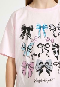 Lyserød bomulds t-shirt med forskellige sløjfedesign i blå, sort, hvid og lyserød samt et trykt slogan "Feeling bow-tiful" nederst.