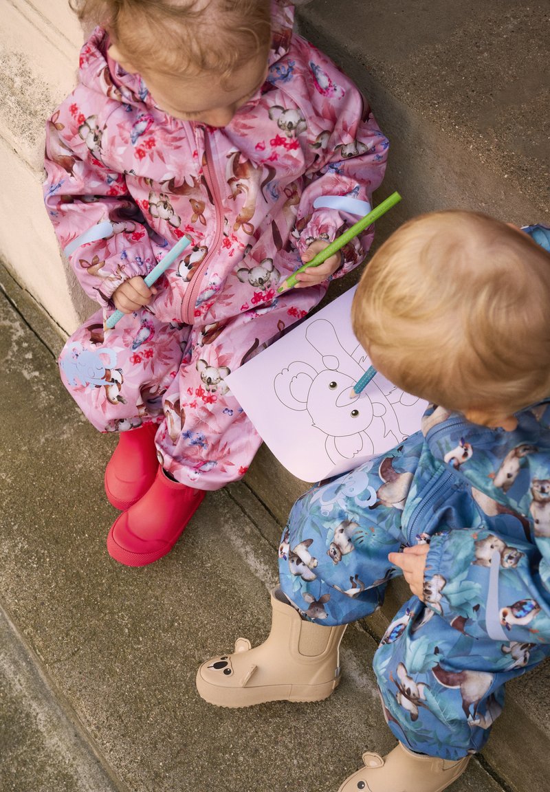 Två barn sitter på betongen. Det ena har på sig en rosa blommig overall med röda stövlar; det andra har på sig en blå overall med djurmönster och bruna björnstövlar.