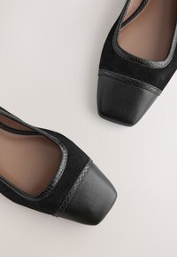 Paire de ballerines noires pour femmes avec bouts carrés en cuir, dessus en daim et bordure texturée sur fond clair.